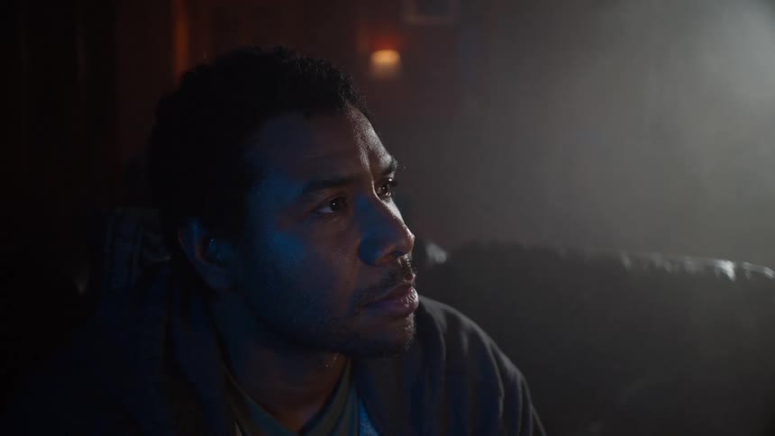 A man watches something off-screen with an intense and focused expression in a dark and hazy room