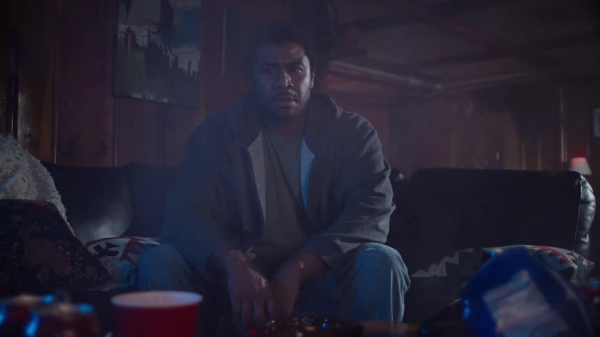 A man loses a video game and sits defeated on his couch in a dark, smoky, and messy room