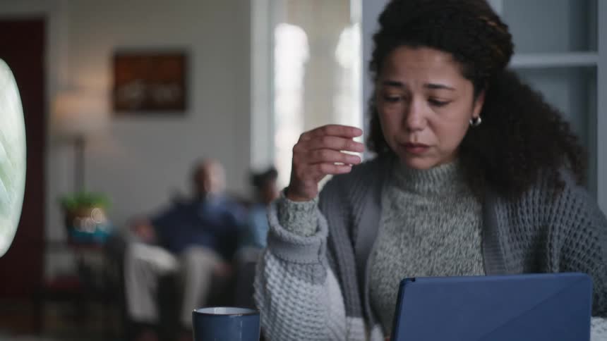 A worried mother deals with financial stress at her dining table while her father and young child play happily in the background