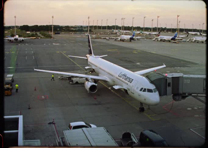 Vintage film shot of a Lufthansa airplane parked at the terminal gate of a busy airport at dusk
