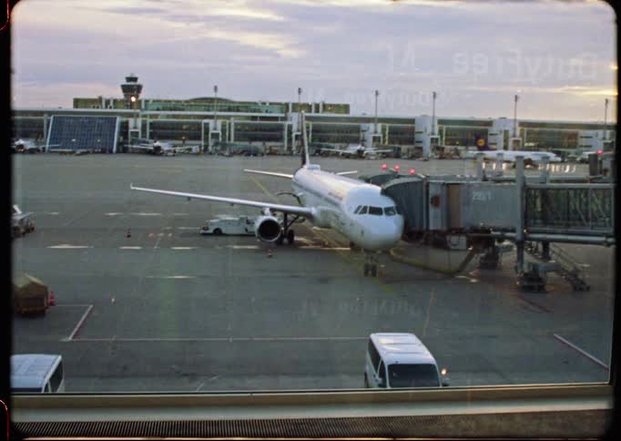 A vintage film-style shot zooms out to reveal a passenger airplane docked at an airport terminal gate