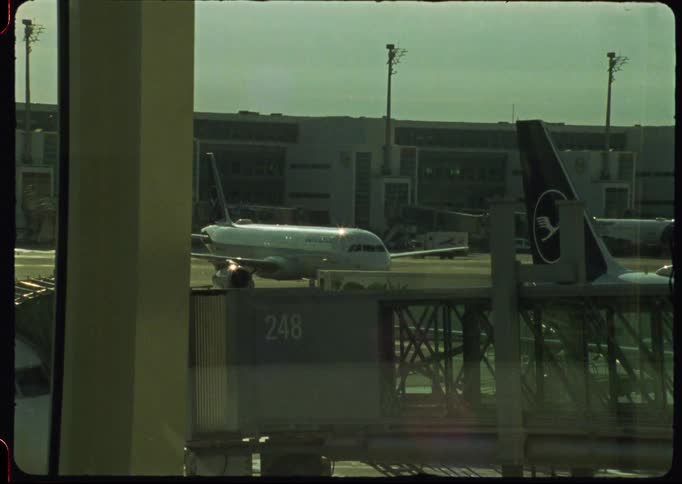 Vintage 16mm analog footage captures a Lufthansa airplane taxiing away from the gate at a bustling airport terminal