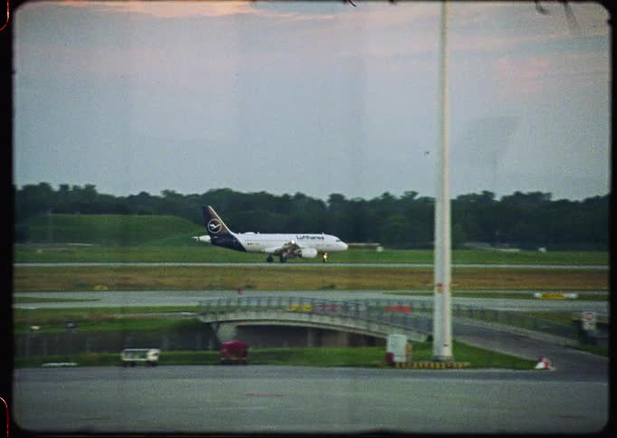 A vintage 8mm film captures a Lufthansa airplane taxiing down the runway on an overcast day