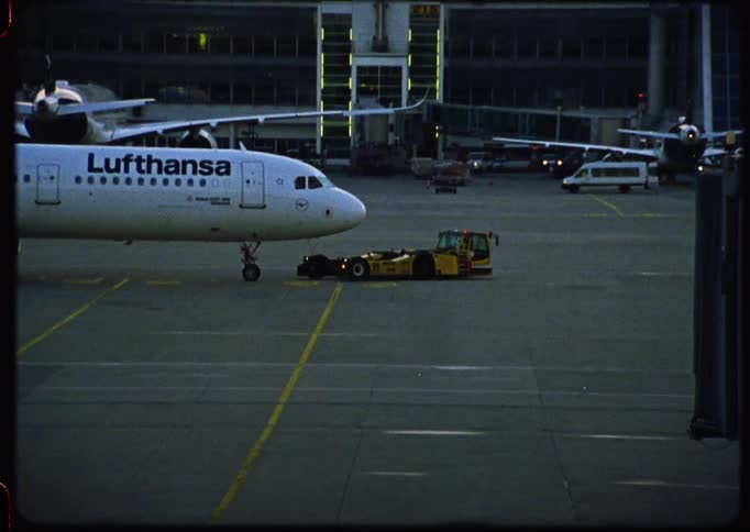 A vintage Super 8mm film captures a Lufthansa airplane on the tarmac at dusk with the airport terminal and control tower in the background