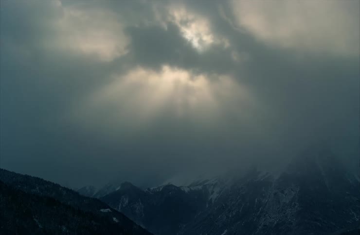 Dramatic sun rays break through dark, moody clouds over a snow-covered mountain range