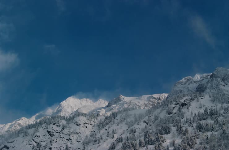 Majestic snow-covered mountains stand tall against a deep blue sky as wispy clouds and mist drift across the peaks