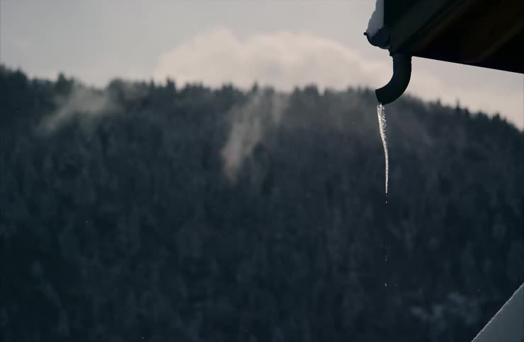 A close-up slow-motion shot captures an icicle melting and dripping from a roof gutter against a backdrop of a misty, snow-covered forest