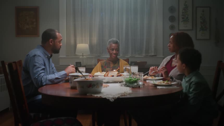 A three-generation African American family gathers for a quiet dinner, with the grandmother sitting at the head of the table