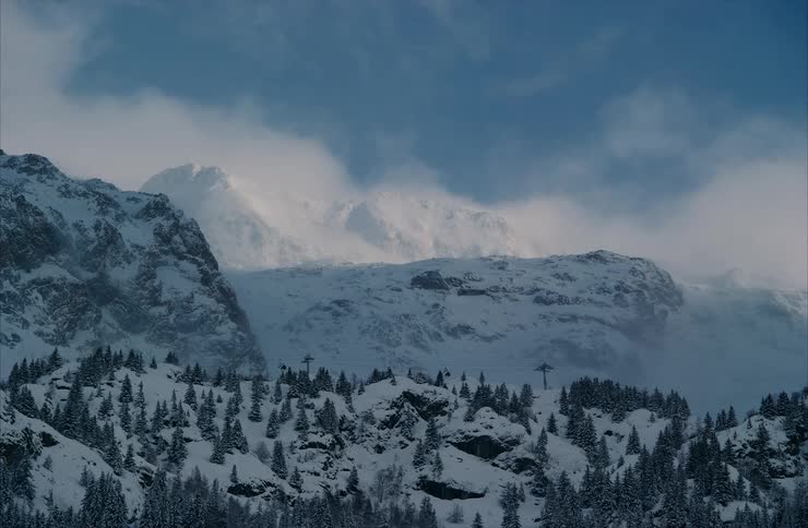 A majestic, static wide shot of a snow-covered mountain range with low clouds moving slowly across the blue sky