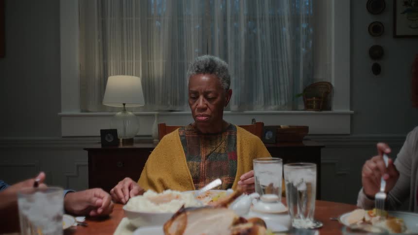 An elderly Black matriarch appears sad and lost in thought during a quiet family dinner with her children and grandchild