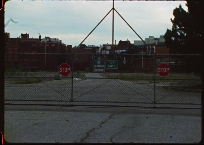 Vintage footage shows the closed chain-link gate entrance to an industrial factory complex on Eastman Avenue