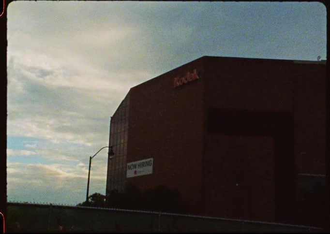 A vintage, grainy film captures a low-angle static shot of the Kodak building with a 'Now Hiring' sign under an overcast sky