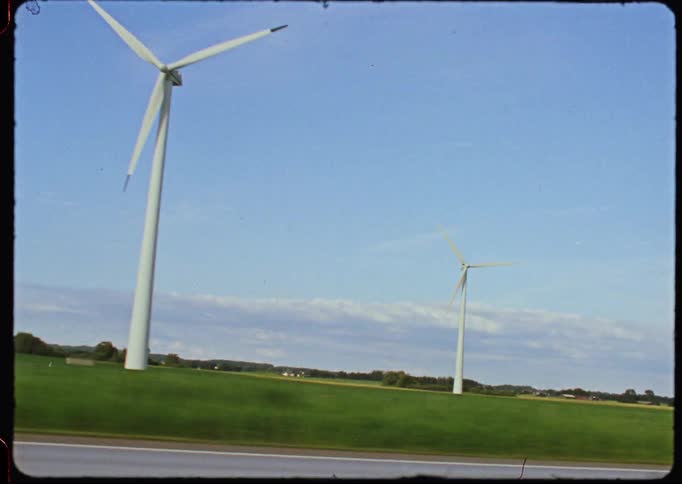 Vintage super 8 style shot from a car driving past wind turbines in a green field under a clear blue sky