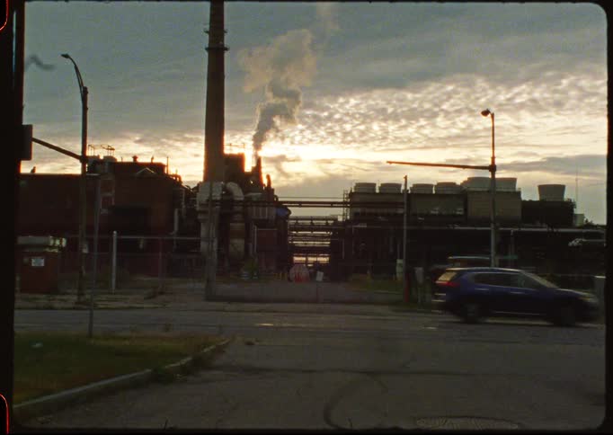 Vintage film captures traffic moving past a sprawling industrial complex with smoke billowing from a smokestack at dawn