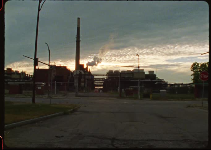Wide shot of a gritty industrial factory with a smokestack releasing smoke as the sun sets on an empty street