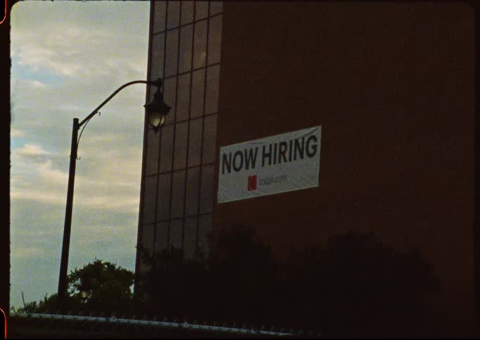 A grainy, vintage shot of a 'Now Hiring' sign hanging on the side of a Kodak building at dusk