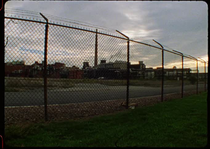 A static film-style shot of an industrial factory with a smoking chimney seen through a barbed-wire fence on an overcast day