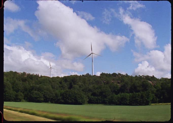 A vintage film-style view from a moving vehicle captures wind turbines spinning on a lush green hill under a partly cloudy sky