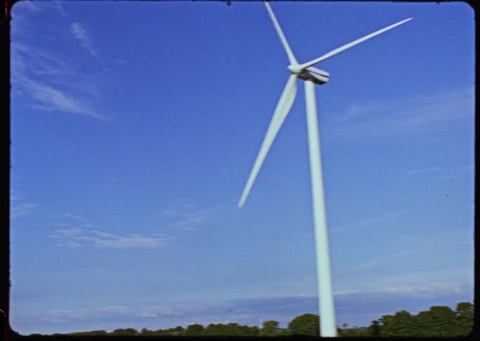 Driving past a large, stationary wind turbine in a rural field under a clear blue sky, with motion-blurred cars and trucks passing in the foreground