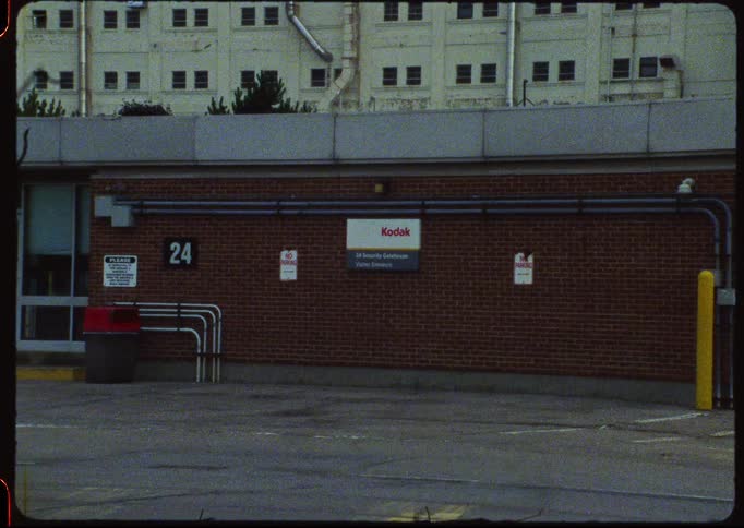 A grainy, vintage shot captures the empty Kodak security gatehouse and visitor entrance