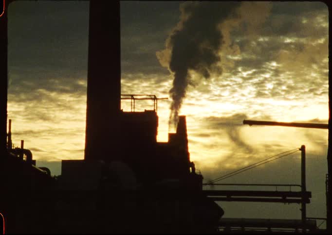 Dark smoke billows from a factory smokestack silhouetted against a dramatic golden sky at dusk