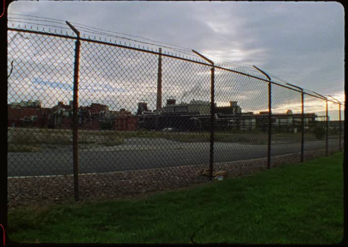 An industrial factory complex with smoking stacks is viewed from behind a barbed wire chain-link fence in this grainy, vintage-style shot