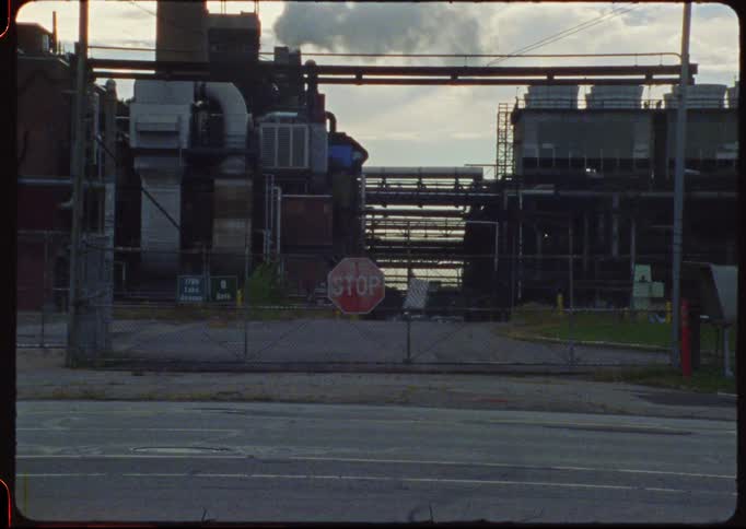 A static vintage shot of a factory entrance blocked by a gate with a stop sign, as smoke rises from a chimney in the background