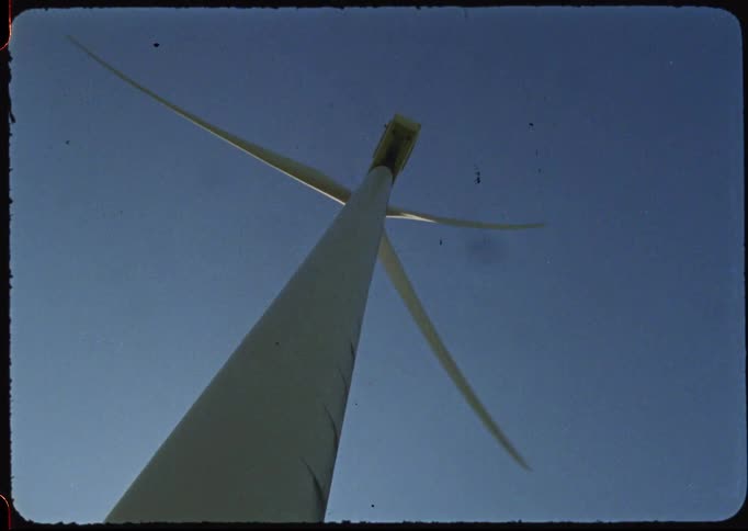 A vintage film shot tilts up a massive wind turbine as its blades spin against the clear blue sky