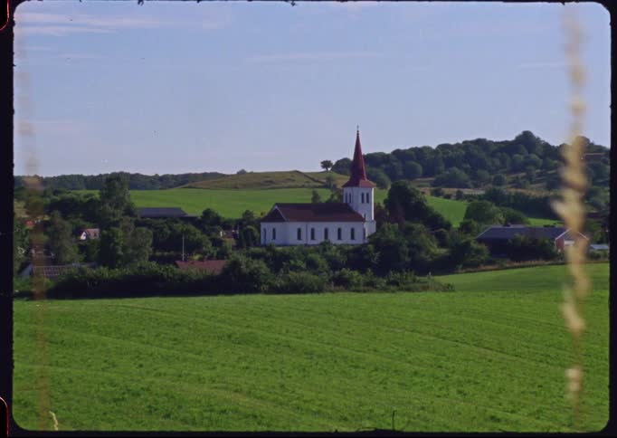 Vintage Super 8 film captures a picturesque white church with a red steeple in a beautiful, green rural landscape