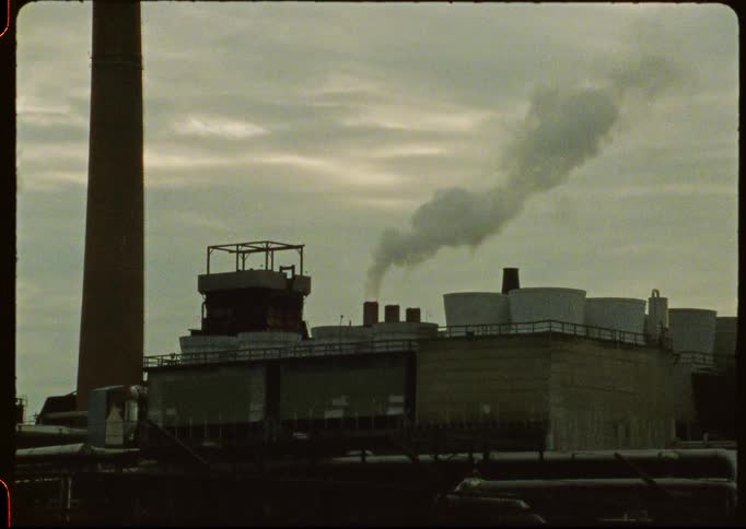 An old industrial factory lets out a plume of smoke from its chimney under a cloudy sky in this vintage footage