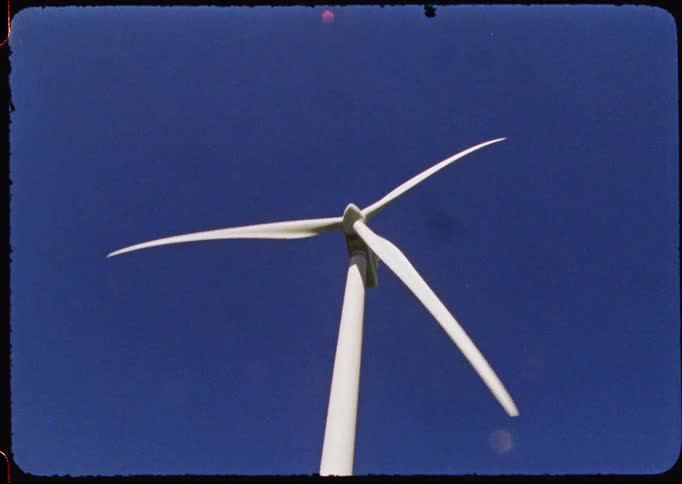 Vintage film shot tilts up a large wind turbine in a grassy field to its spinning blades against a clear blue sky