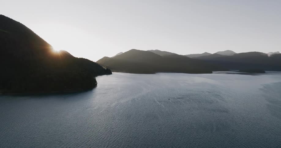 An expansive aerial shot captures the early morning sun rising from behind silhouetted mountains over a tranquil body of water