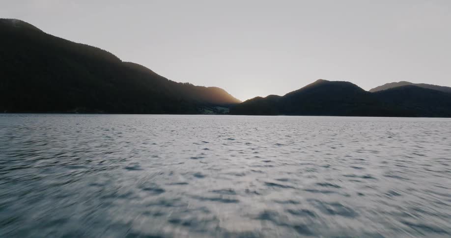A fast aerial shot flies low over the rippling surface of a vast lake towards silhouetted mountains at sunrise
