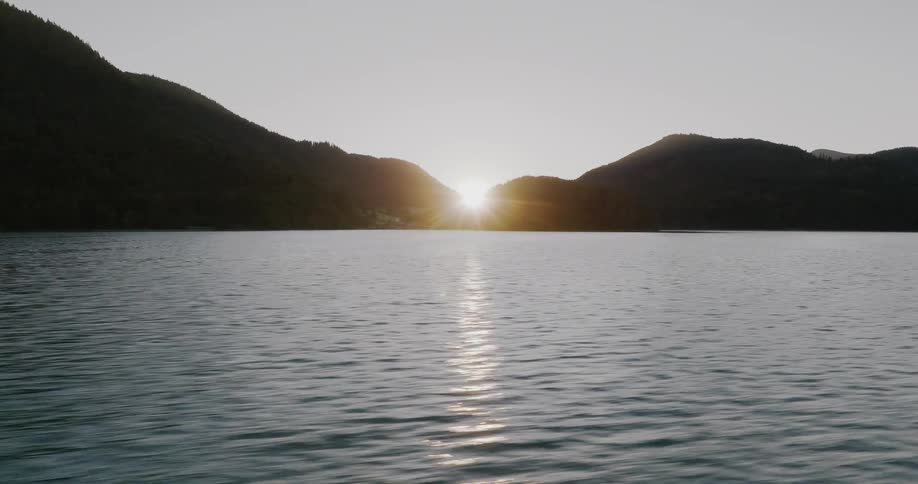 A serene timelapse of the sun rising from behind silhouetted mountains, its golden light creating a starburst and reflecting on the calm lake surface