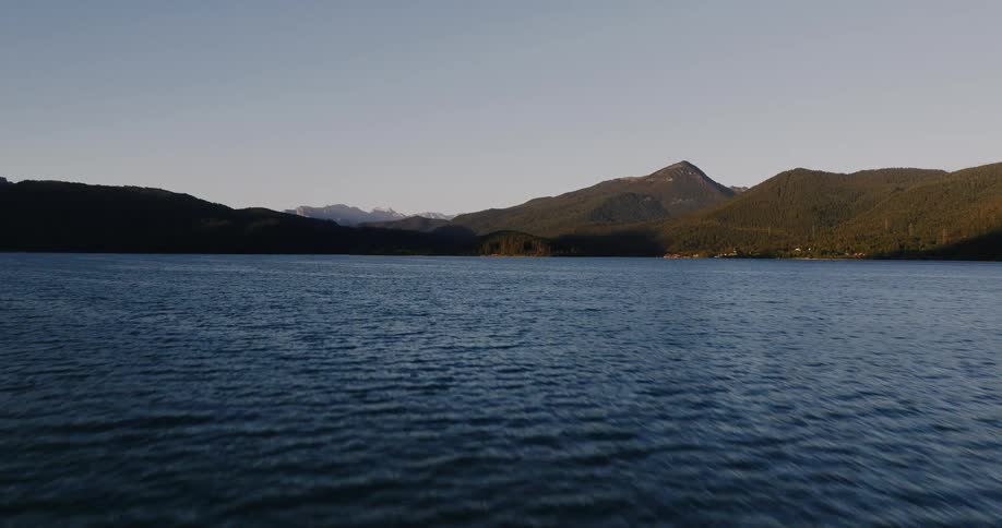 A low aerial shot flies over a serene mountain lake at sunrise, gradually rising to reveal the vast and beautiful landscape