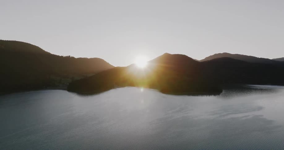 The sun slowly crests over a silhouetted mountain range, its golden light reflecting on the vast, tranquil lake below in a stunning aerial view