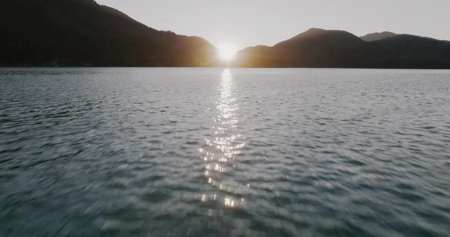Low aerial pullback over gentle water ripples reveals a beautiful sunset between silhouetted mountains