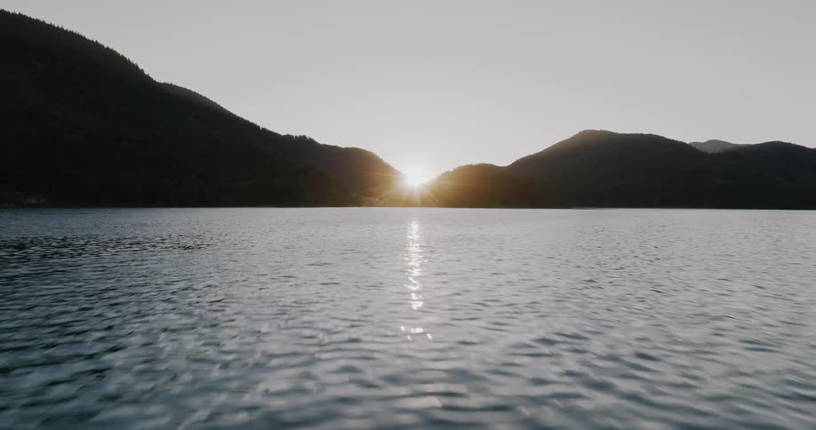An exhilarating low-altitude drone shot glides quickly across a serene lake as the sun sets between silhouetted mountain peaks