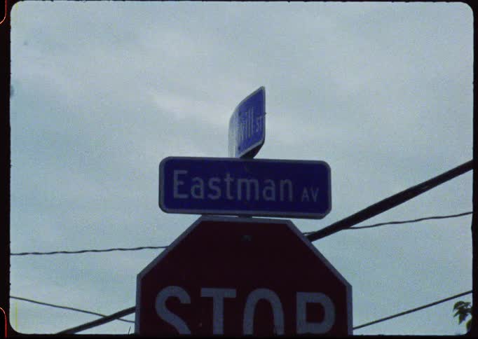 A vintage, grainy film shot of an 'Eastman AV' street sign and a stop sign against a cloudy, overcast sky