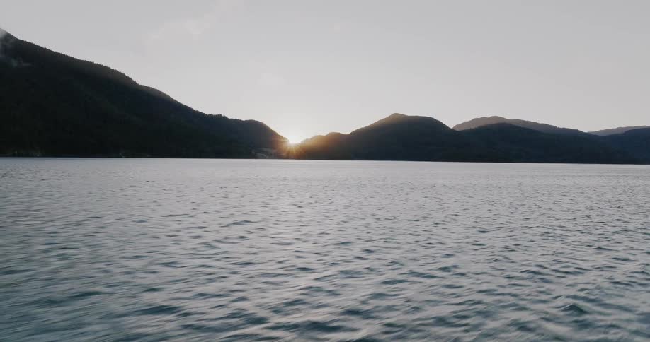 A serene timelapse captures the sun rising over silhouetted mountains, its reflection shimmering on the calm surface of a vast lake