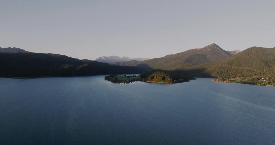 A majestic aerial panorama of a calm alpine lake with a forested island and surrounding mountains at sunrise