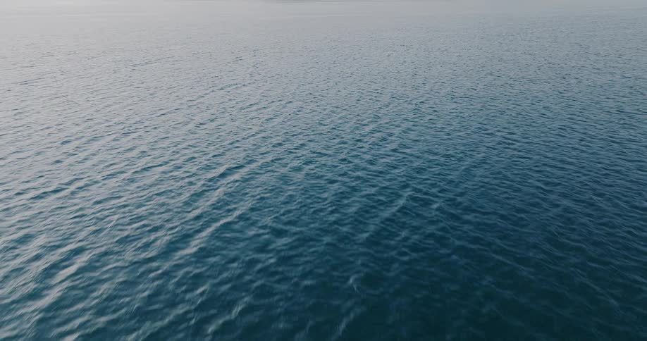 An aerial view flying over the calm, deep blue sea with gentle ripples stretching to the horizon