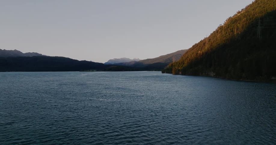 An aerial view flies over a vast mountain lake as the warm sunset light hits the forested hillsides