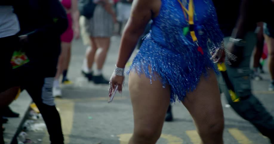 A low-angle slow-motion shot of people dancing in the street during a carnival, focusing on a woman in a sparkly blue costume