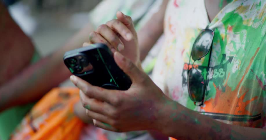 A close-up of a young woman's paint-splattered hands as she scrolls on her smartphone during a color festival