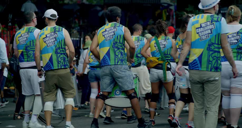 A colorful samba drumming band wearing matching uniforms marches down the street in a slow-motion parade