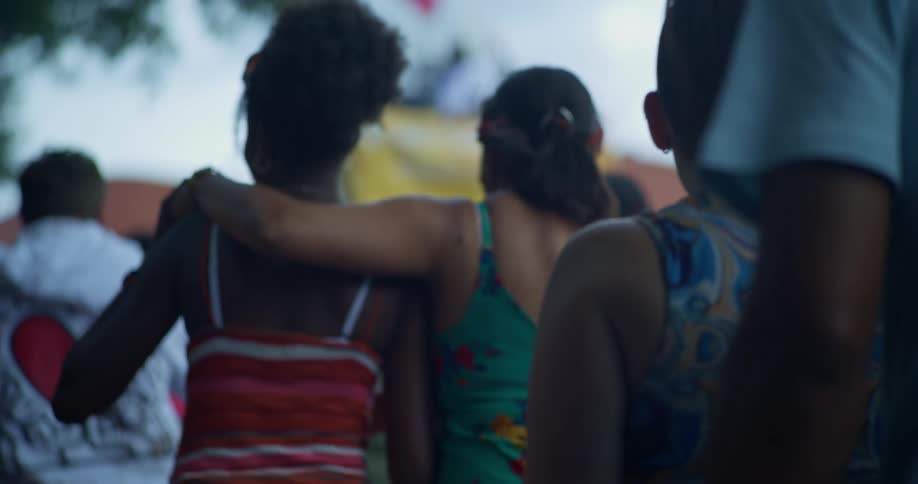 Two women walk arm in arm through a diverse crowd at an outdoor summer festival in slow motion