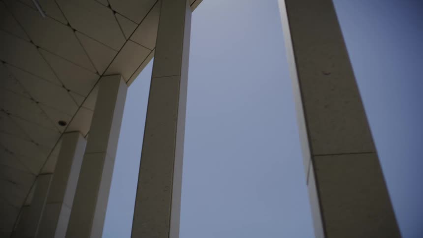 A low-angle slow pull-out reveals the grand scale of modern architectural columns against a cloudless blue sky