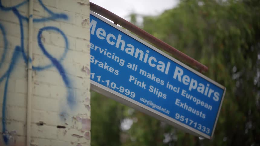 A blue 'Mechanical Repairs' sign hangs next to a graffiti-covered brick wall on an overcast day