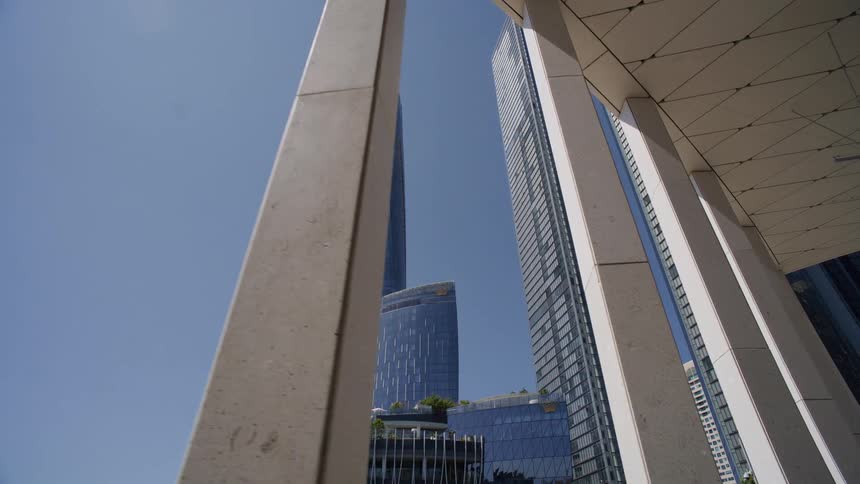 A slow dolly shot reveals modern glass skyscrapers framed between architectural pillars on a bright, sunny day